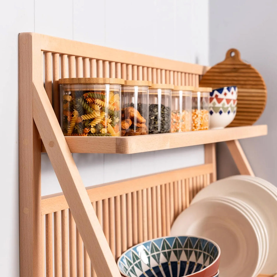 A side view of a plate shelf kitchen rack showing the detail of the wood. Food is being store in jars along the top shelf and a large bowl sits in the fore ground. Plates line the back. 