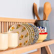 Detailed view of the top shelf of a plate rack. Cups, saucers and bowls are on display. In the distance there is a blue enamel utensil holder with wooden spoons sat upright.